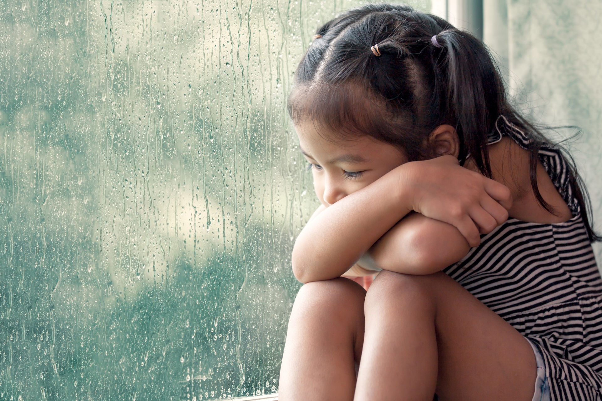 A young girl sits by a window on a rainy day, hugging her knees and looking down thoughtfully. Raindrops are visible on the glass, and she appears quiet and reflective.