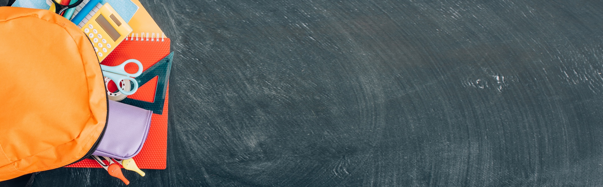An orange backpack with school supplies like notebooks, scissors, pens, a calculator, and a pencil case spilling out onto a dark chalkboard background with empty space on the right.
