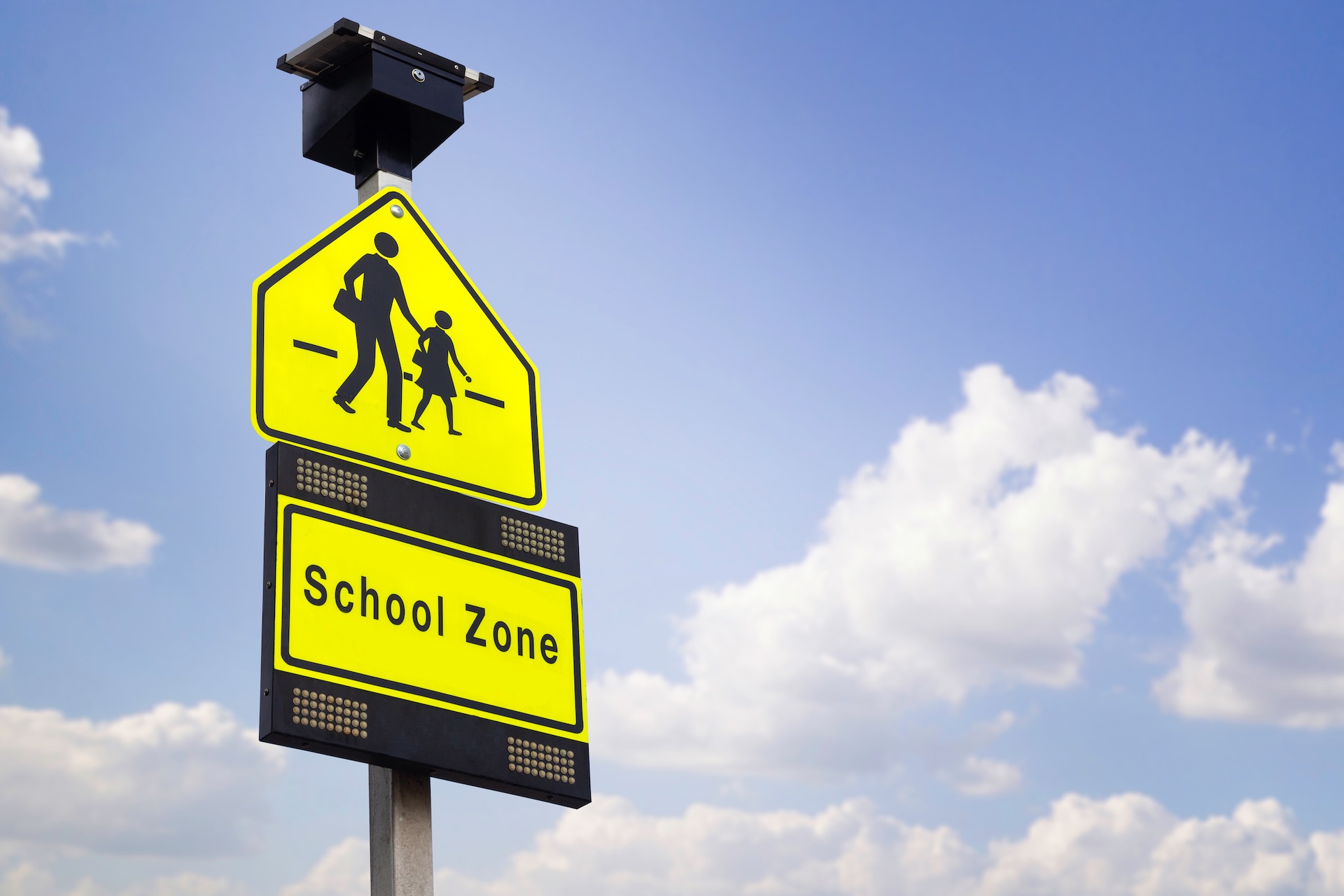 Bright yellow school zone sign with an image of an adult and child walking, topped with a small solar panel, set against a partly cloudy blue sky.