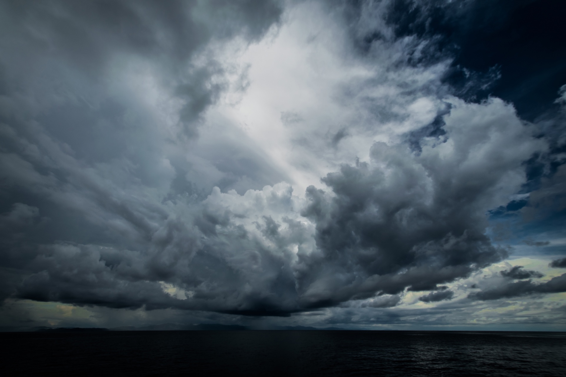 Dramatic dark clouds gather over a calm sea, creating a moody atmosphere with patches of light breaking through the stormy sky.