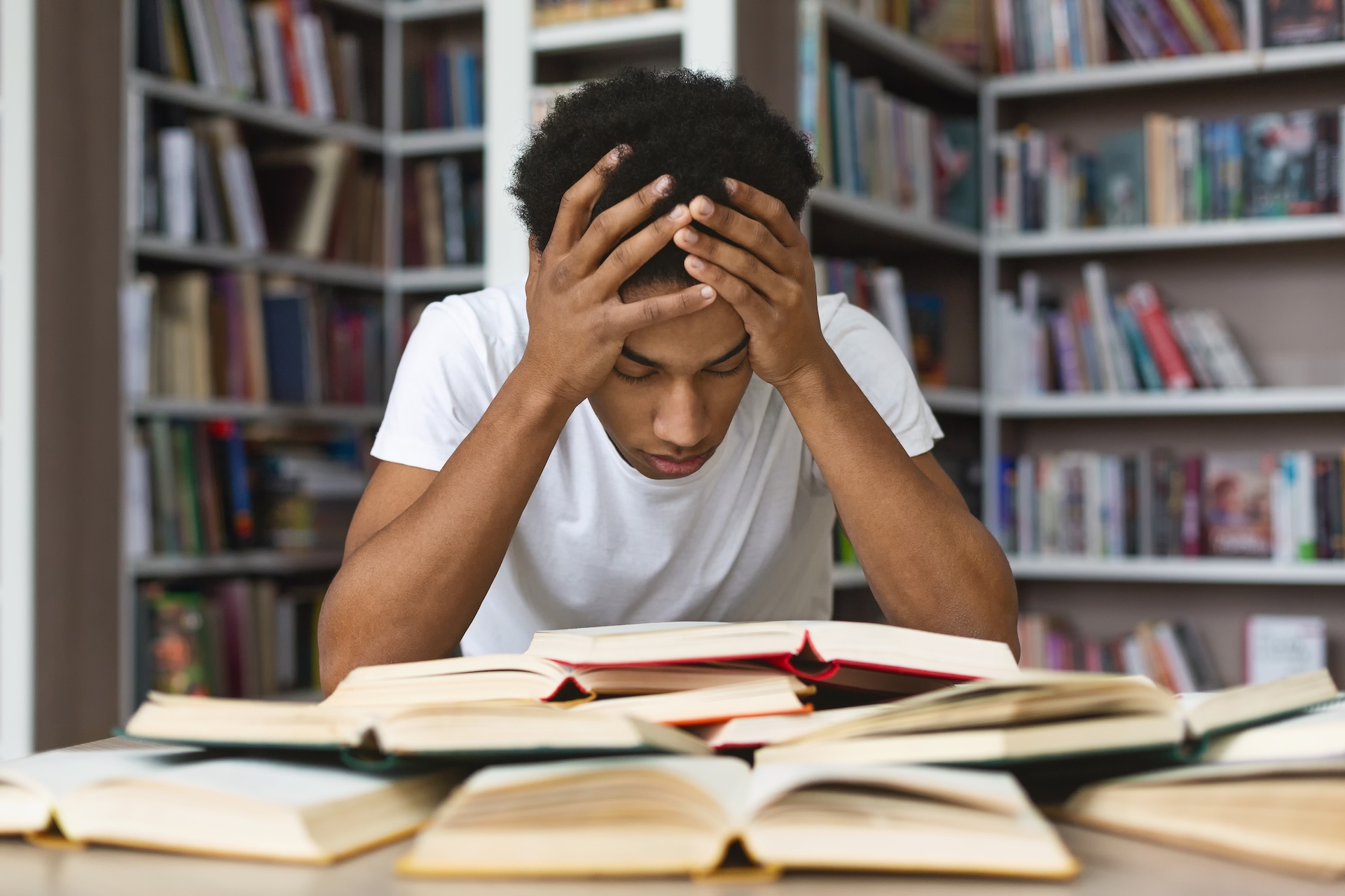A person sits at a desk in a library, holding their head in their hands and looking down at several open books, appearing stressed or overwhelmed by studying. Shelves filled with books are in the background.