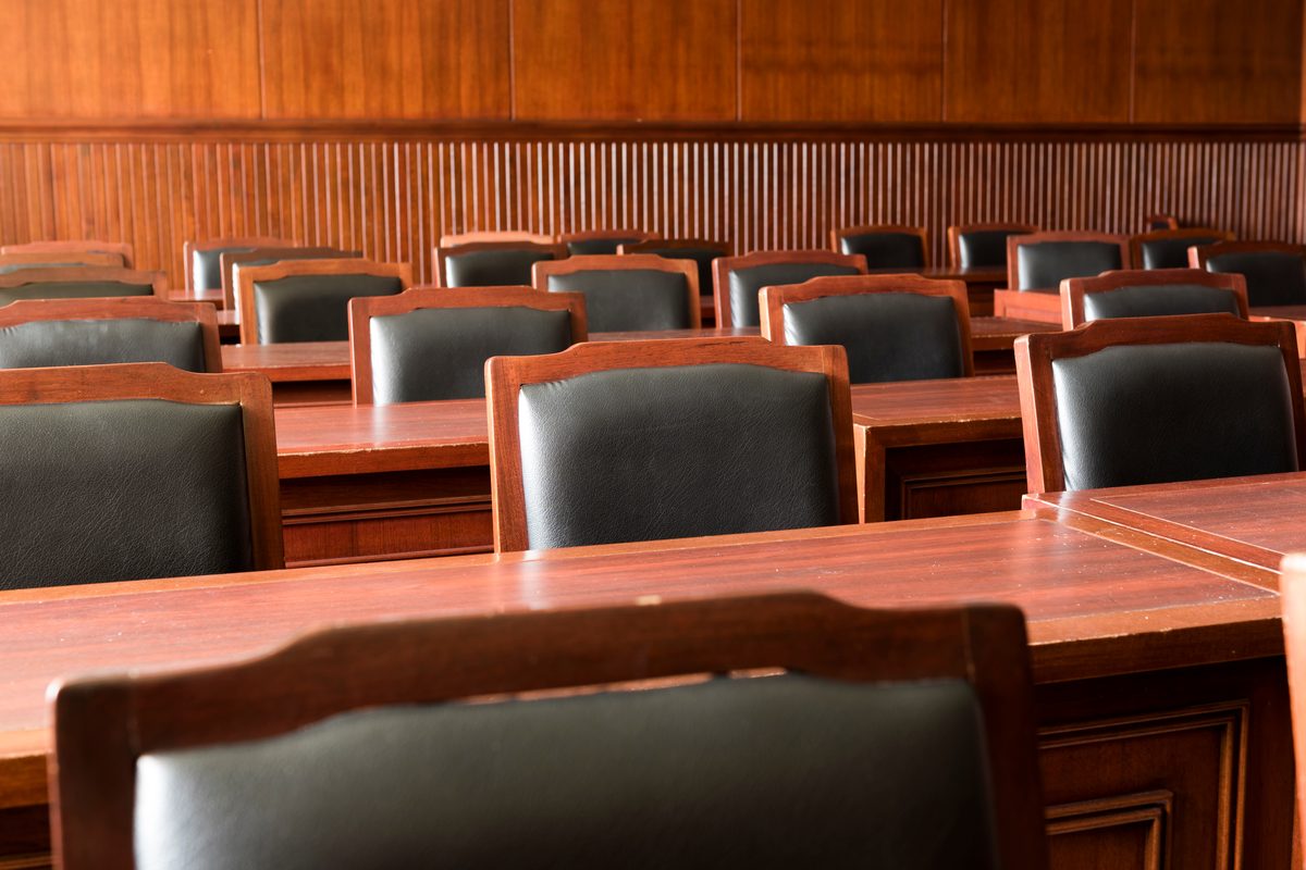 Rows of empty wooden benches and chairs inside a quiet courtroom. Rows of empty wooden benches and chairs inside a quiet courtroom.