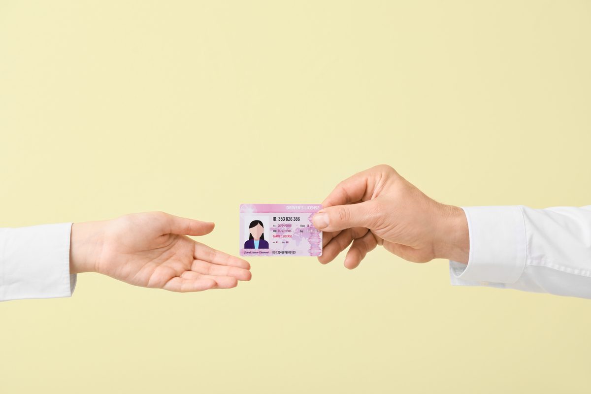 One person handing a driver license card to another person's open palm against a plain yellow background. One person handing a driver license card to another person's open palm against a plain yellow background.