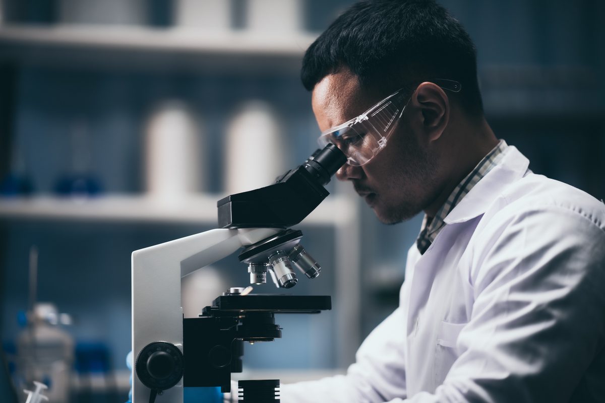 Forensic scientist in a white lab coat and safety glasses examining a sample through a microscope in a darkened laboratory. Forensic scientist in a white lab coat and safety glasses examining a sample through a microscope in a darkened laboratory.