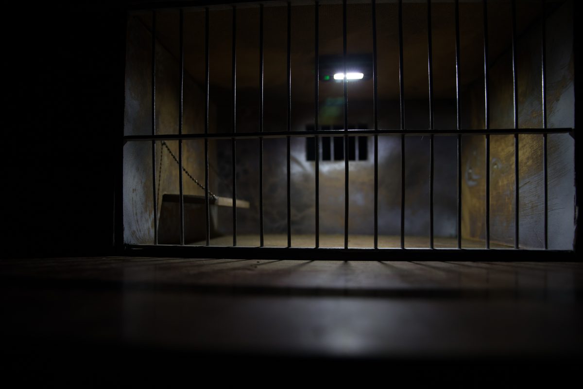 The interior of an empty jail cell seen through steel bars under dim overhead lighting. The interior of an empty jail cell seen through steel bars under dim overhead lighting.