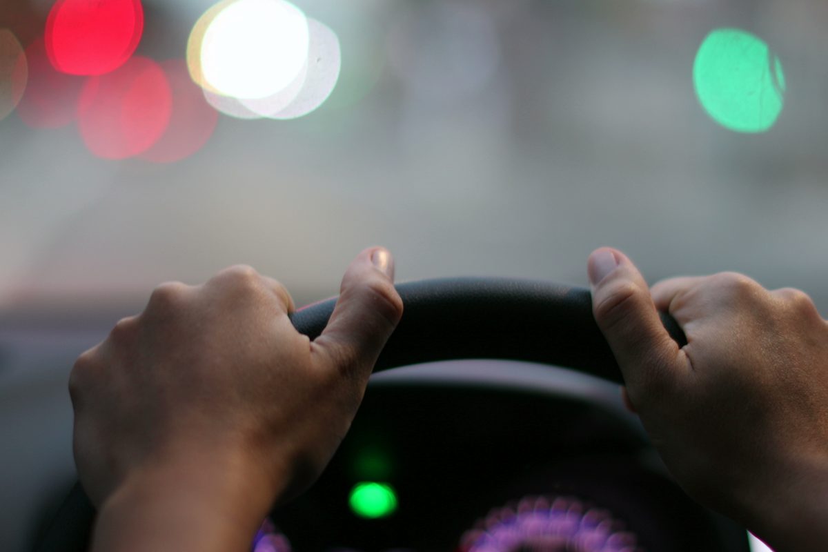 Driver's point of view of two hands resting on a steering wheel at night, with city lights blurred through the windshield.