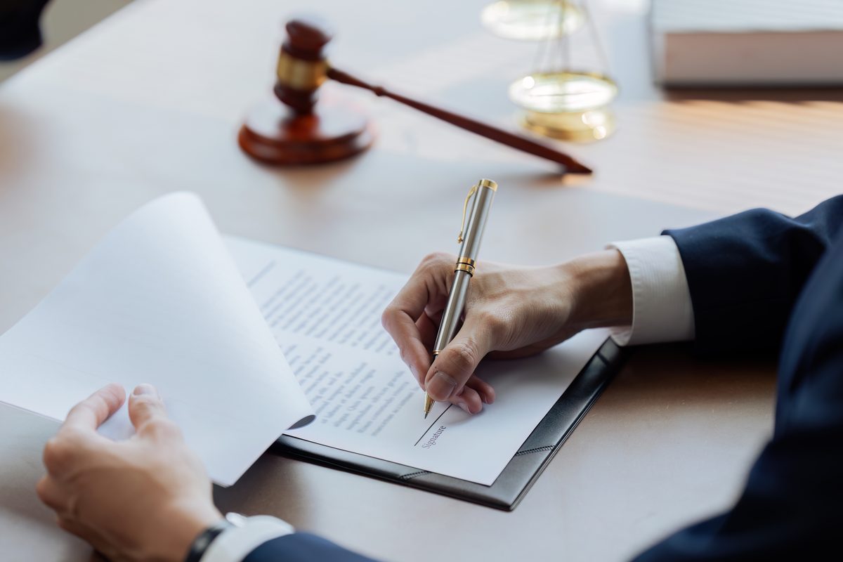 A person in a dark suit signing a legal agreement with a fountain pen, with a wooden judge's gavel and scales of justice on the desk. A person in a dark suit signing a legal agreement with a fountain pen, with a wooden judge's gavel and scales of justice on the desk.