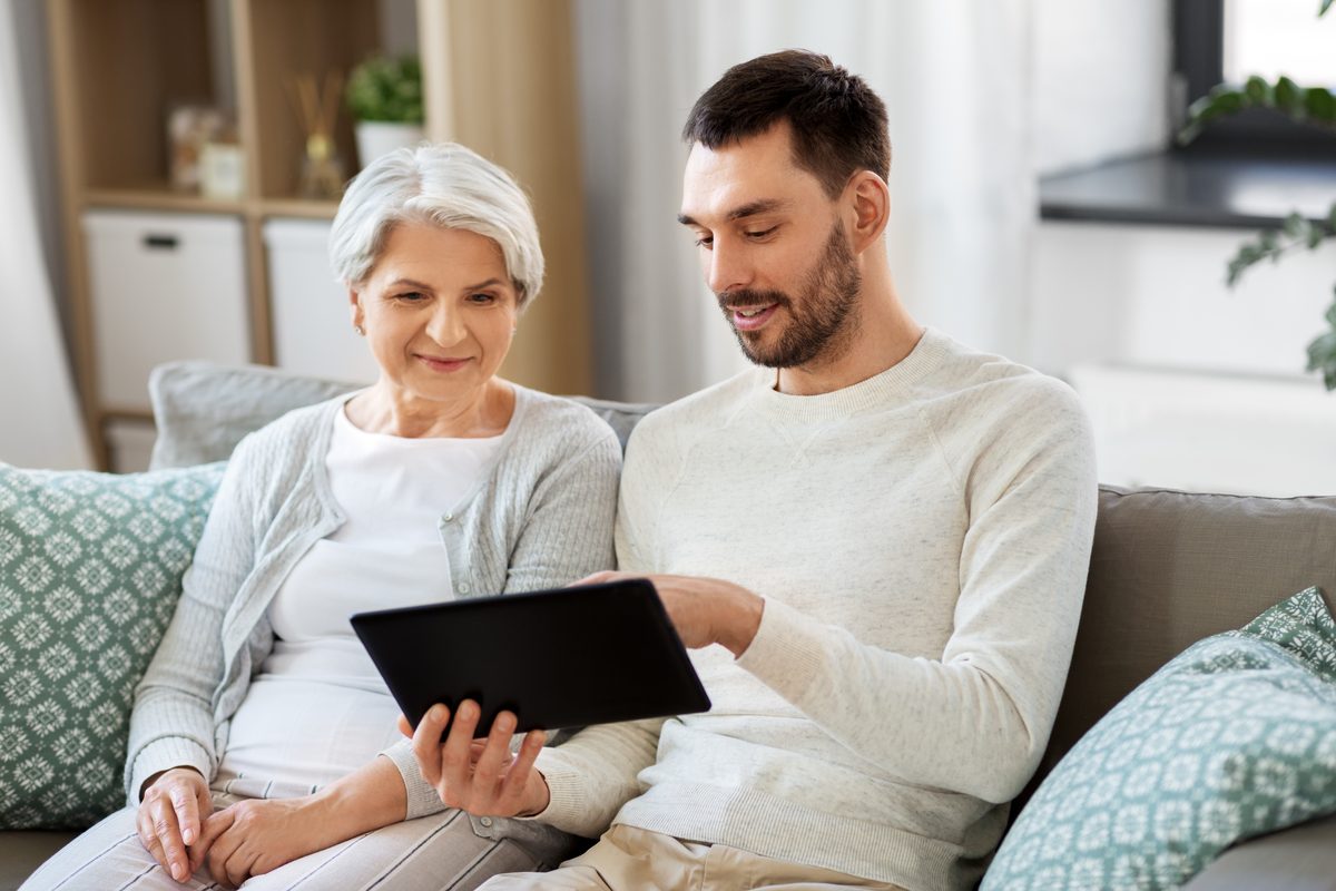 An older woman and a younger man sitting together on a sofa looking at a tablet. An older woman and a younger man sitting together on a sofa looking at a tablet.